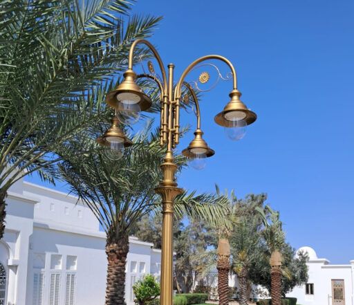 Lampadaire richement décoré avec un abat-jour doré et quatre luminaires 04/Aries/A, sur fond de palmiers et de bâtiments blancs par une journée ensoleillée.