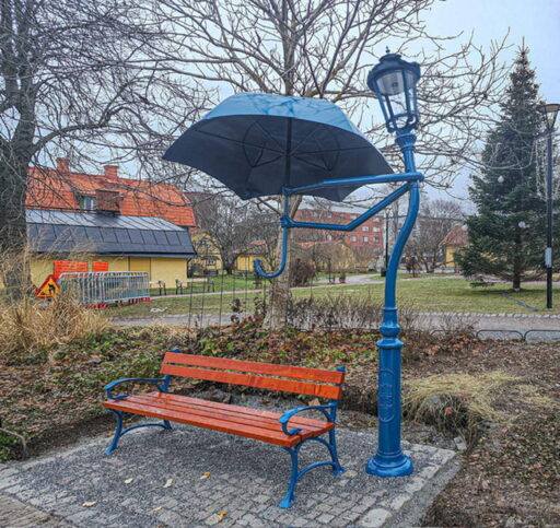 Une lampe de rue bleue en forme de personnage tenant un parapluie au-dessus d'un banc dans un parc.